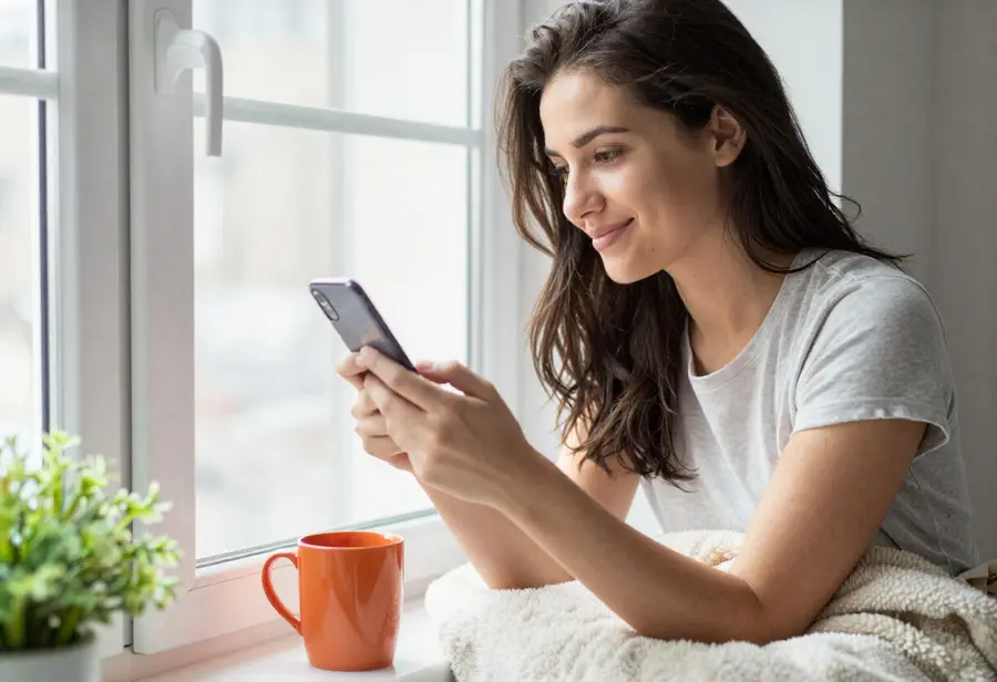 A woman sitting on a window seat with a blanket, smiling quietly at her phone